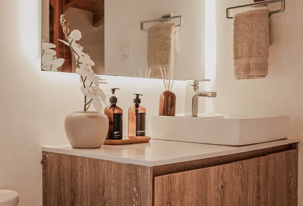 Ein Badezimmer mit einem weißen Waschbecken, einem Spiegel und einem Holzkabinent. Auf der Ablage stehen zwei Seifenflaschen und eine Vase mit weißen Blumen.