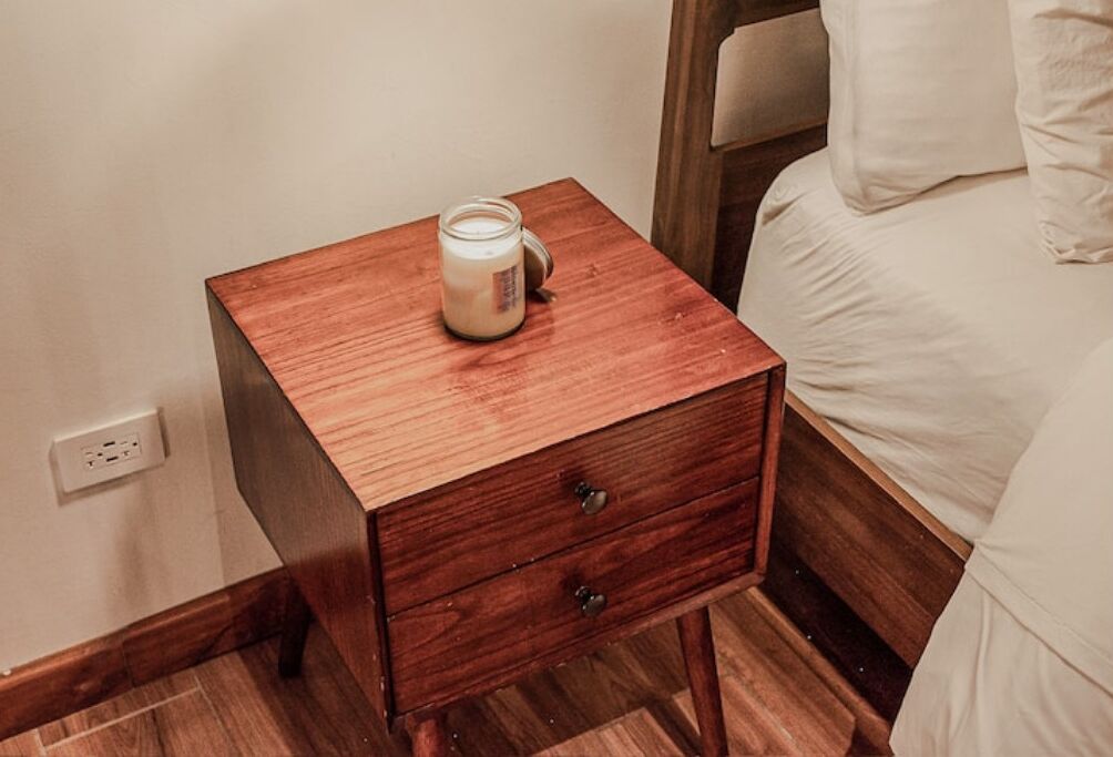 Een houten nachtkastje met een potje room naast een bed met wit beddengoed.