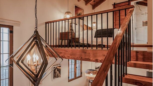 Una escalera de madera con barandilla de metal que conduce a un dormitorio en altillo con una cama y una lámpara colgante.