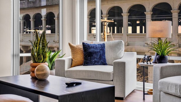 A modern living room with a grey sofa, a coffee table, and a lamp. There are potted plants and a view of a historic building with arches. - City Getaway, Free Parking, 24/7 Hour Gym (Cleveland)