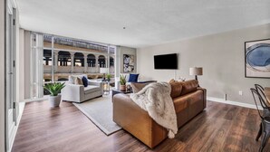 A modern living room with a brown leather sofa, a white armchair, and a coffee table. There is a large window with a view of a bridge and buildings. A flat-screen TV is mounted on the wall, and there are two potted plants on the floor. - City Getaway, Free Parking, 24/7 Hour Gym (Cleveland)