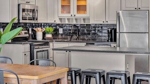 A modern kitchen with white cabinets, a stainless steel refrigerator, a black tiled backsplash, and a wooden dining table with bar stools. - City Getaway, Free Parking, 24/7 Hour Gym (Cleveland)