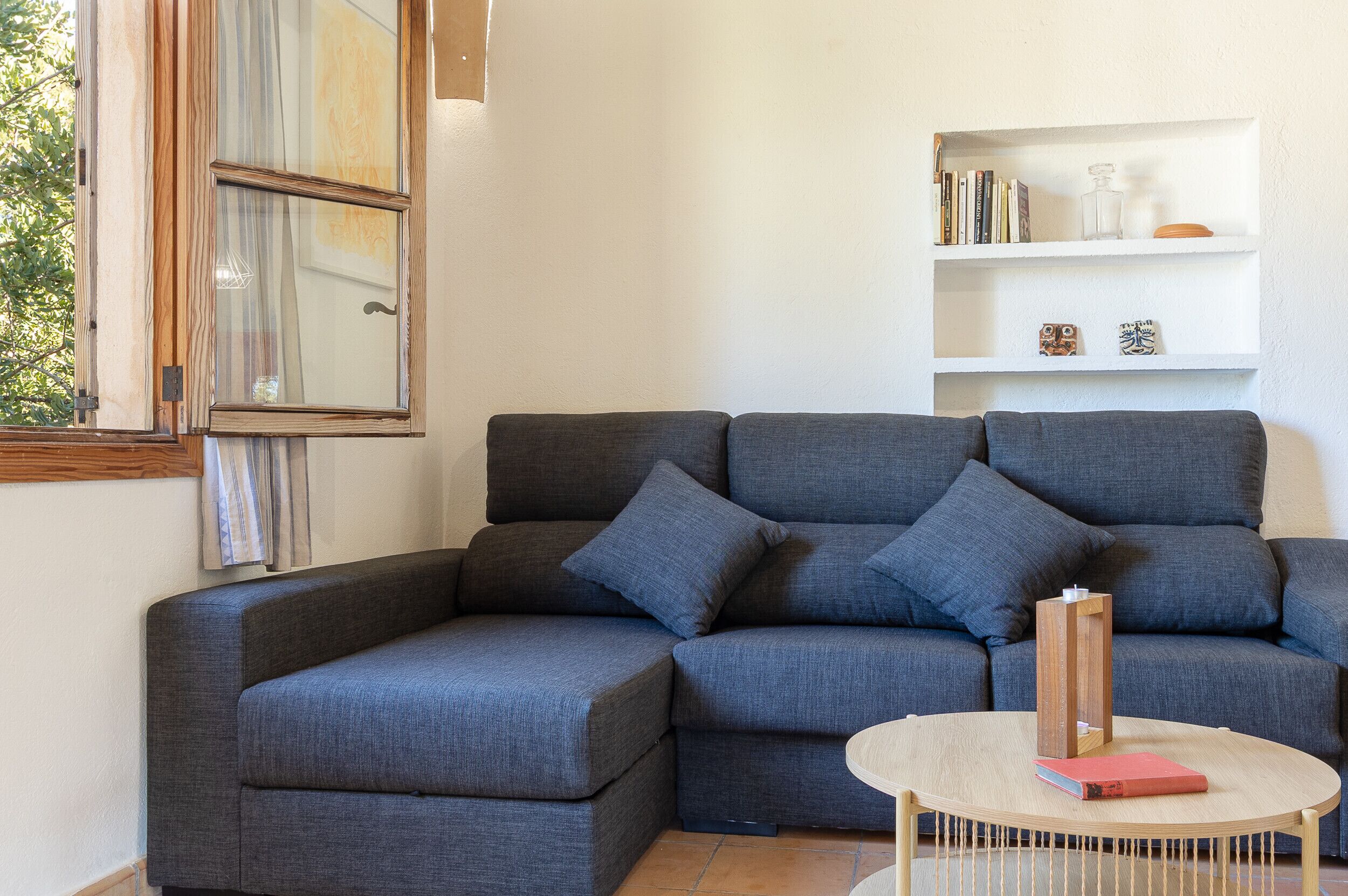 A corner sofa with cushions, a small round table with a book, and a white shelf with decorative items.