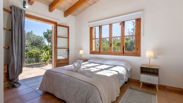 Une chambre à coucher avec un grand lit, deux tables de chevet, une fenêtre avec des cadres en bois et une vue sur la verdure.