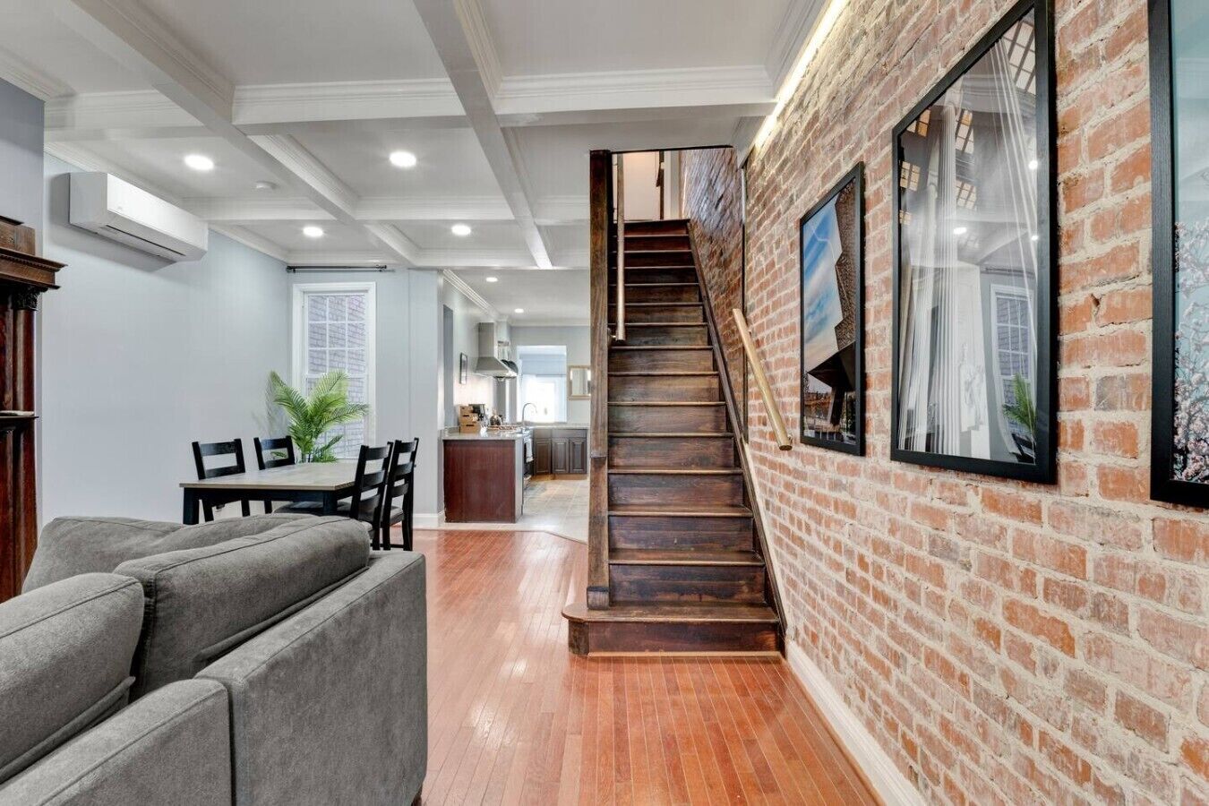A modern interior with a staircase leading to a kitchen area, a dining area with a gray sofa, and a brick wall with framed artwork.