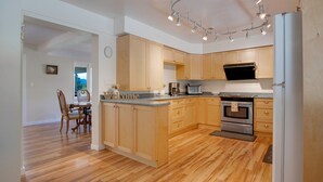 A modern kitchen with wooden cabinets, a central island, stainless steel appliances, and a dining area with a chair and table. - Oceanfront Getaway with Private Dock + Beach (Sooke)