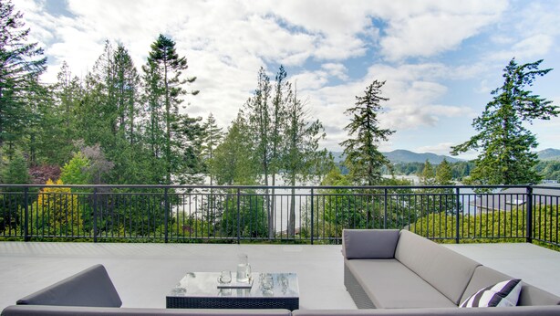A balcony with a sofa and a table, overlooking a forested area with mountains in the distance. - Oceanfront Getaway with Private Dock + Beach (Sooke)
