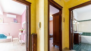 Una stanza con un muro rosa, una porta in legno, una panca bianca, un tavolino con una pianta e un bagno con pavimento piastrellato di verde.