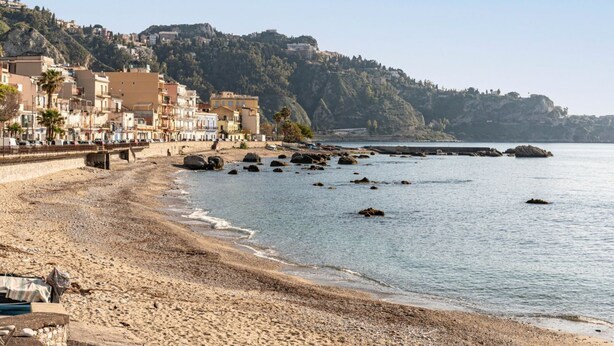Una località balneare con edifici lungo la spiaggia, un litorale roccioso e una collina con strutture sullo sfondo.