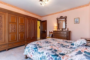 A bedroom with a floral bedspread, a wooden wardrobe, a dresser, and a mirror on the wall.