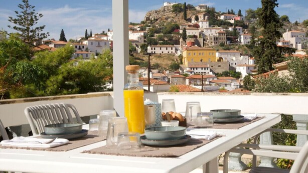 A terrace with a table set for breakfast, overlooking a hillside town with buildings and greenery. - Elegant Hydra Getaway (Idra)