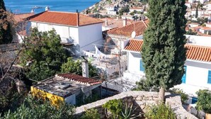 A coastal town with terracotta-roofed buildings, a prominent cypress tree, and a patio with chairs and potted plants. - Historical Stone Manor-Upper Floor (Idra)