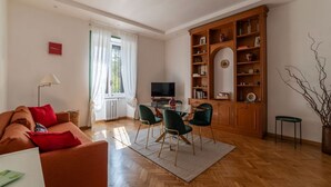 Een woonkamer met een bank, stoelen, een televisie, een boekenkast en een raam met gordijnen.