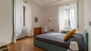 Een slaapkamer met een bed, een stoel, een klein tafeltje, een radiator en een raam met gordijnen.