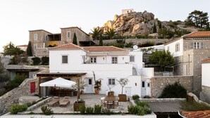 A traditional stone building with a white facade, a patio with outdoor furniture, and a view of rocky hills in the background. - Villa Alethea Retreat (Idra)