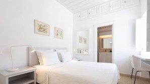 A white bedroom with a bed, bedside tables, a desk, and chairs. There are two framed pictures on the wall. - Villa Alethea Retreat (Idra)