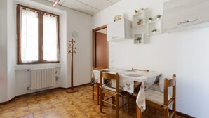 Een eetruimte met een tafel, stoelen, een radiator, een raam met gordijnen, een klok en wandplanken met planten.
