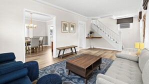 Un vaste salon comprenant une table basse en bois, un canapé bleu et un canapé gris. Un escalier mène à l’étage supérieur, et on y trouve également un coin repas agrémenté d’un lustre.