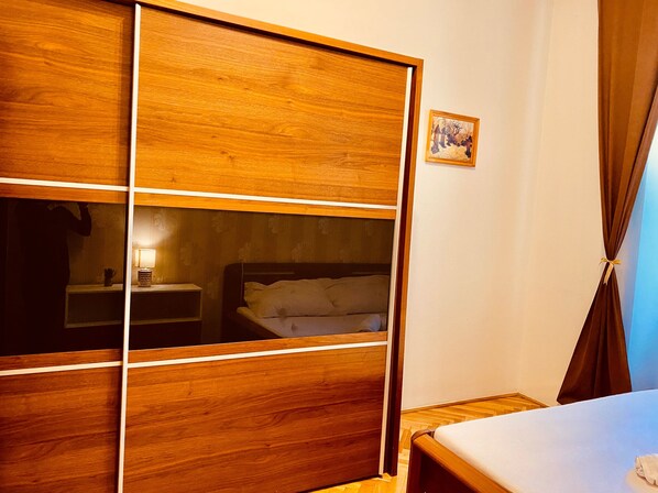 A room with a wooden wardrobe, a bed, a nightstand, and a small table with a lamp.