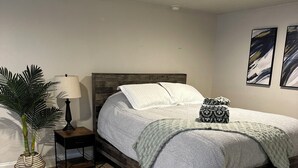 Une chambre à coucher avec un cadre de lit en bois, une table de chevet, une lampe, une plante en pot et des œuvres d’art abstraites accrochées au mur.