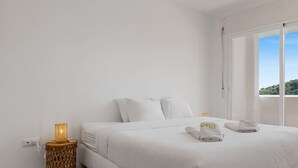 A white bed with pillows, a bedside table with a lamp, and a window with a view of greenery. - Rosa | Sea views - pool - terrace (Mijas Costa)