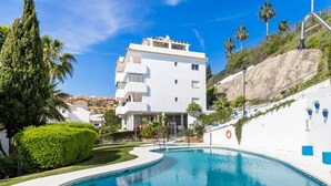 A swimming pool with a curved edge, surrounded by a white building with balconies, a grassy area, and palm trees. - Rosa | Sea views - pool - terrace (Mijas Costa)