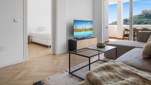 A modern living room with a TV displaying a beach scene, a sofa, a coffee table, and a view of a balcony with a scenic landscape. - Rosa | Sea views - pool - terrace (Mijas Costa)