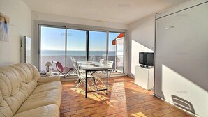 Een moderne woonkamer met een bank, een eettafel en stoelen. Er staat een televisie op een dressoir, en een groot raam met uitzicht op zee.