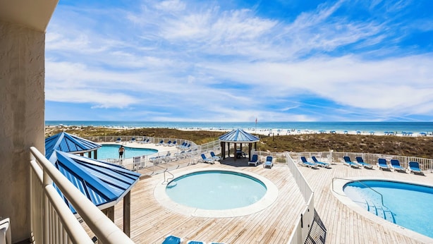 A wooden deck with two pools, lounge chairs, and a gazebo overlooking a beach.