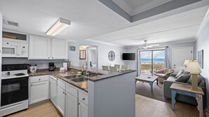 A modern kitchen with white cabinets, a microwave, and a stove. There is a dining area with a table and chairs, and a balcony with a view of the ocean.