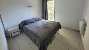 A bedroom with a bed, a nightstand, a radiator, and a window with a view of greenery. - CAP D'AGDE - Plage de la Roquille / Réf 251 (CAP D'AGDE)