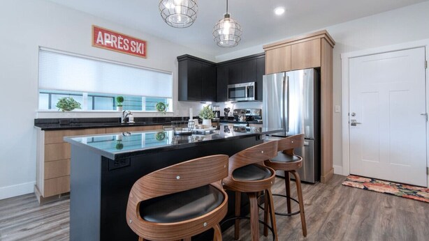 A modern kitchen with a central island, black cabinets, stainless steel appliances, and wooden bar stools. - Modern 2-BR Townhome w/ Garage, AC & Workspace (Driggs)