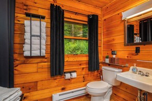 Une salle de bain rustique, aménagée dans une cabane en bois, comprenant des toilettes, un lavabo, un porte-serviettes et une fenêtre avec des rideaux.