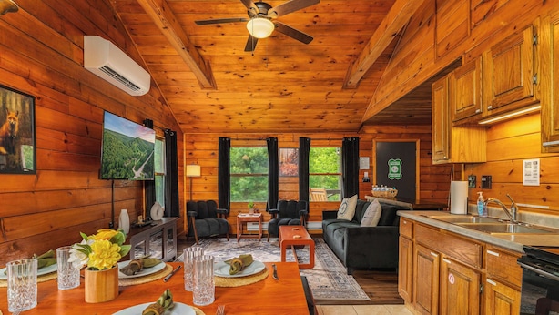 Interior de una cabaña de madera con comedor, sofá, cocina y televisor.