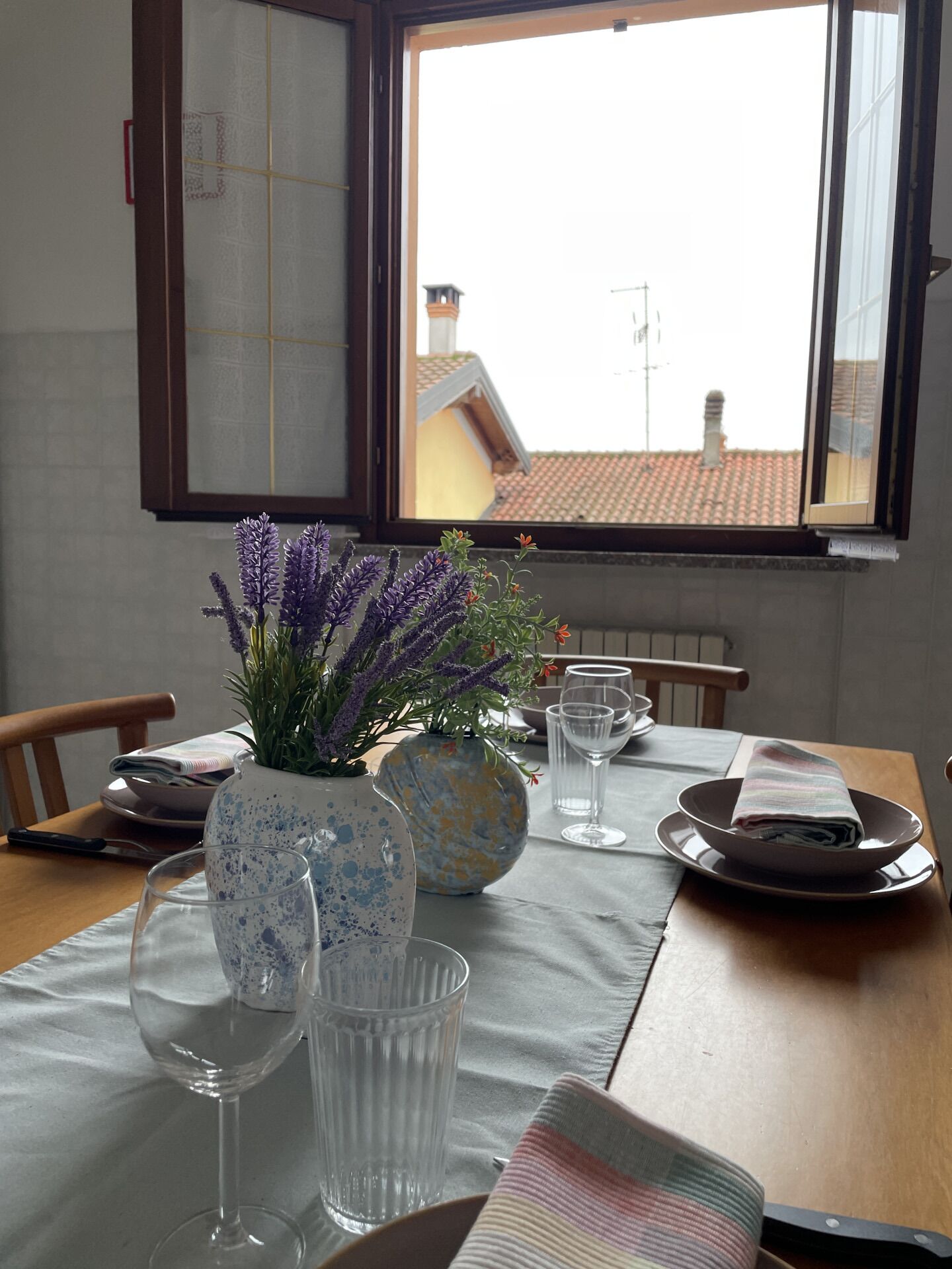 A dining table with a vase of purple flowers, a blue and yellow ceramic pot, and two wine glasses.
