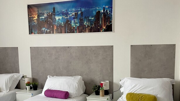 A hotel room with two beds, a cityscape view, and a nightstand with a lamp and a plant. - Bnbook Residenza Vespucci 2 (sedriano)