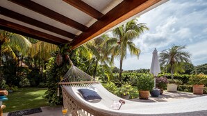 Un hamac suspendu sous une pergola en bois, entouré d’une végétation luxuriante et de plantes en pot.