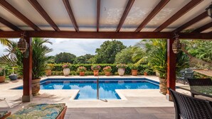 Une piscine extérieure couverte, agrémentée d’une pergola en bois, entourée de plantes en pot et d’un jardin luxuriant.