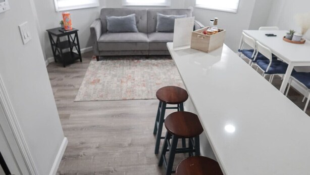 A modern kitchen with a white countertop, two stools, a grey sofa, and a small table with a box and a bottle.