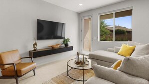 Una sala de estar moderna con sofá, sillón y mesa de centro. Un televisor de pantalla plana está montado en la pared y hay un ventanal corredizo que da a un área exterior.