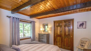 A bedroom with a wooden ceiling, a bed with a striped blanket, a dresser, a chair with a pillow, and a door with a wooden panel. - Loose Moose Lodge – Family chalet, fireplace, ski! (Bartlett)