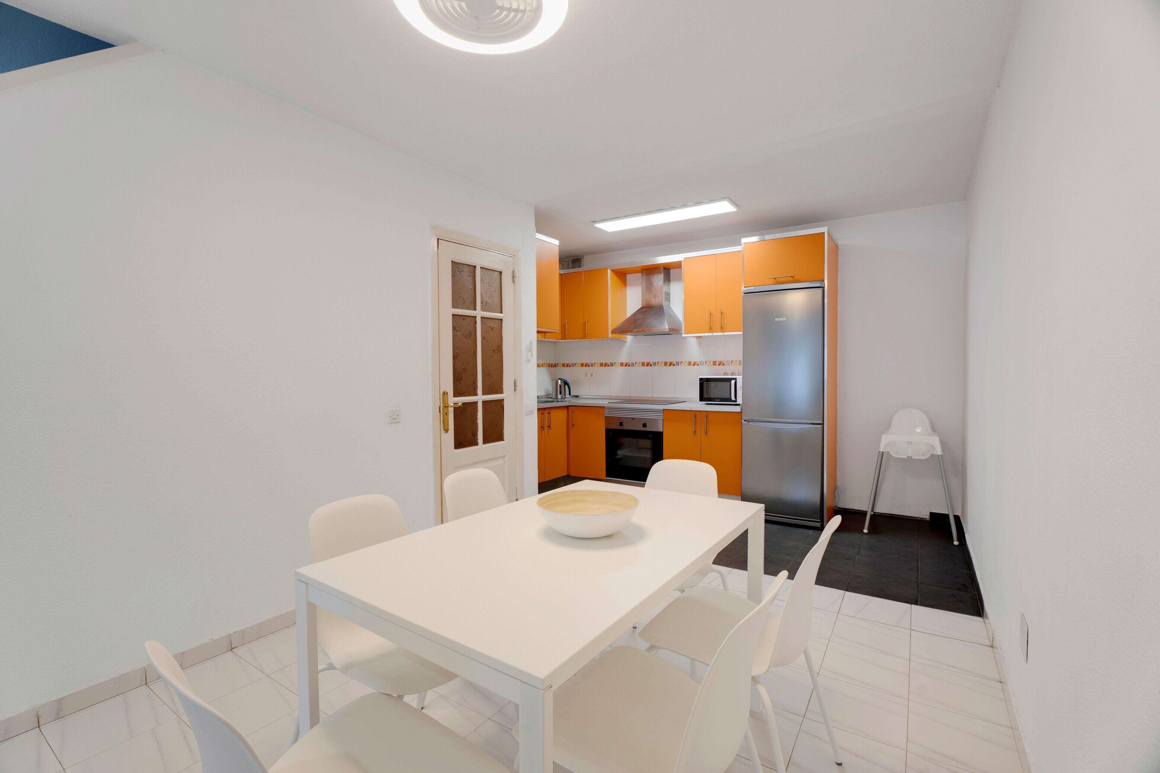 Cocina compacta con electrodomésticos modernos, comedor con mesa y sillas blancas, y una pequeña silla blanca en la esquina.