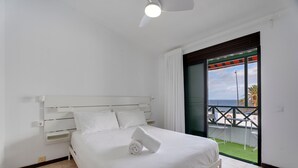 Quarto com cama branca, ventoinha no teto e vista para o mar através de uma varanda.