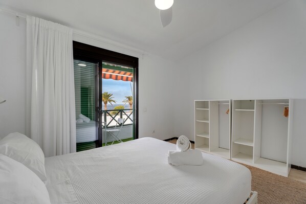 A hotel room with a bed, a wardrobe, a balcony with a view of palm trees, and a window with blinds.