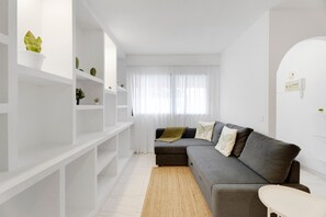 A modern living room with a grey sofa, white shelving, and a large window with sheer curtains.
