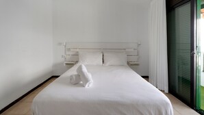 Quarto de hotel com cama, cortinas brancas, porta de correr de vidro e cabeceira branca.