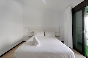 Habitación de hotel con cama, cortinas blancas, puerta corrediza de vidrio y cabecera blanca.