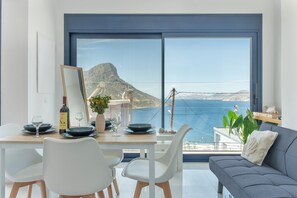 A dining area with a table set for two, a sofa, and a large window with a view of the sea and mountains. - Apartment 'Regina 1' with Sea View, Wi-Fi and Air Conditioning (Kalymnos)