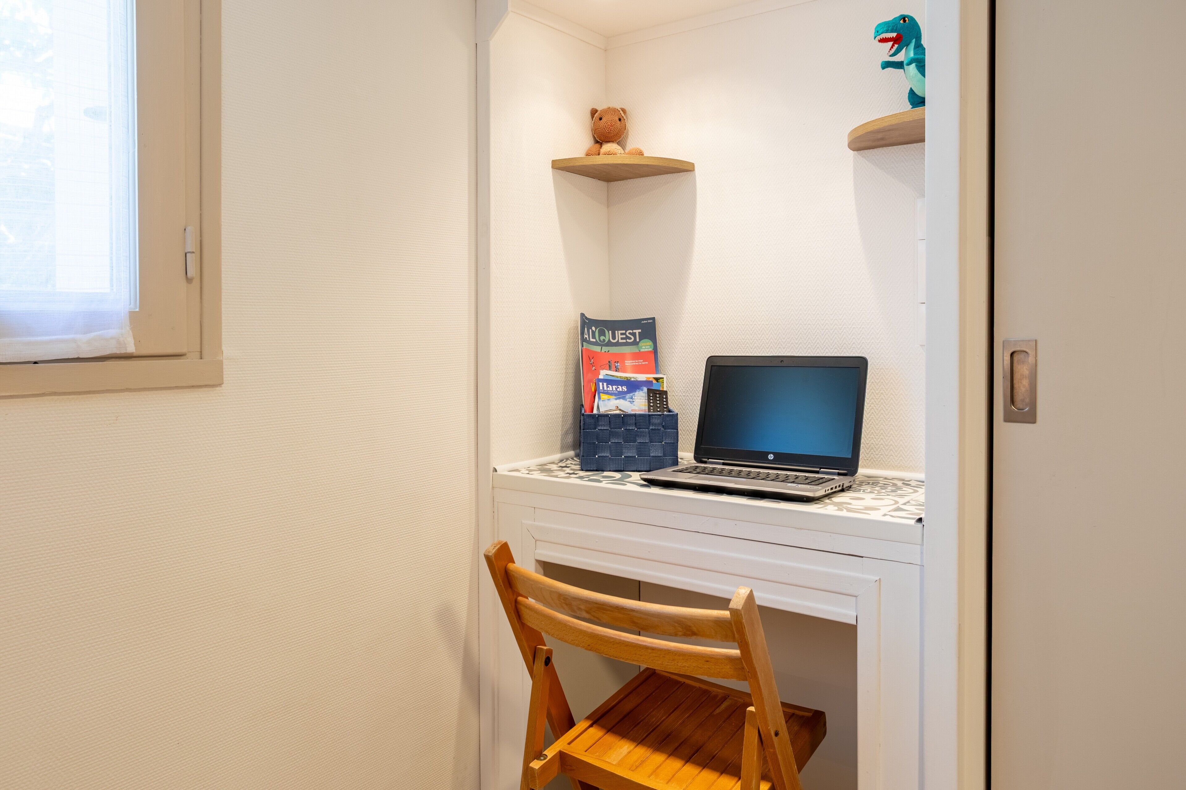 A small room with a desk, a laptop, a chair, a teddy bear on a shelf, and a dinosaur decoration.