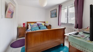A bedroom with a wooden bed, a window with purple curtains, a bedside table with a lamp, and a wall-mounted TV.
